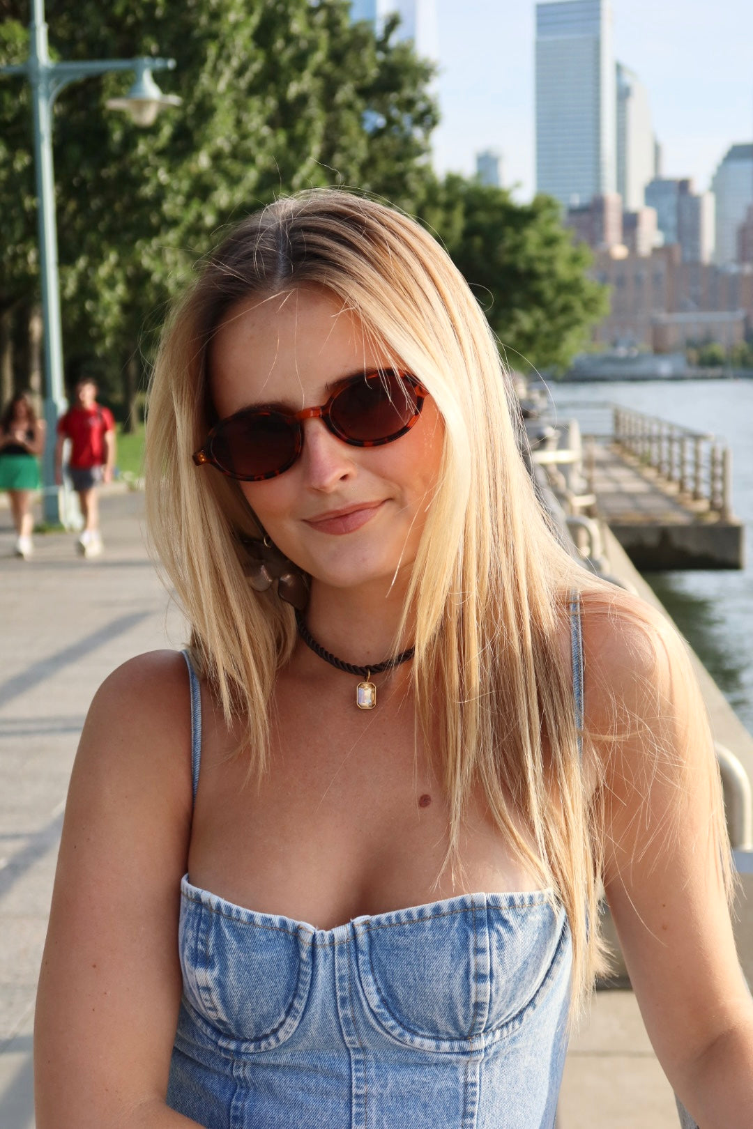 Woman wearing sunglasses and a denim dress by a waterfront with city skyline in the background