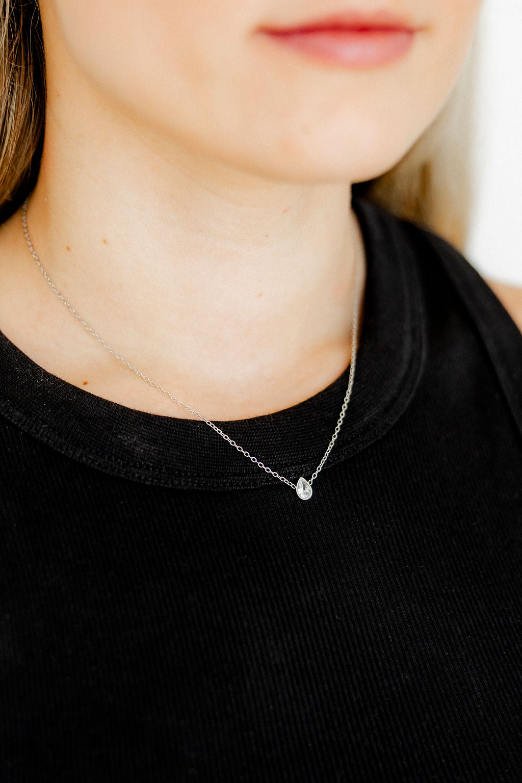 A person wearing a black shirt showcasing a silver necklace with a pear-shaped clear crystal pendant at the center, with the necklace resting neatly around the neck.