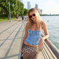 Woman in denim top and leopard print skirt standing by a waterfront with trees and buildings in the background.