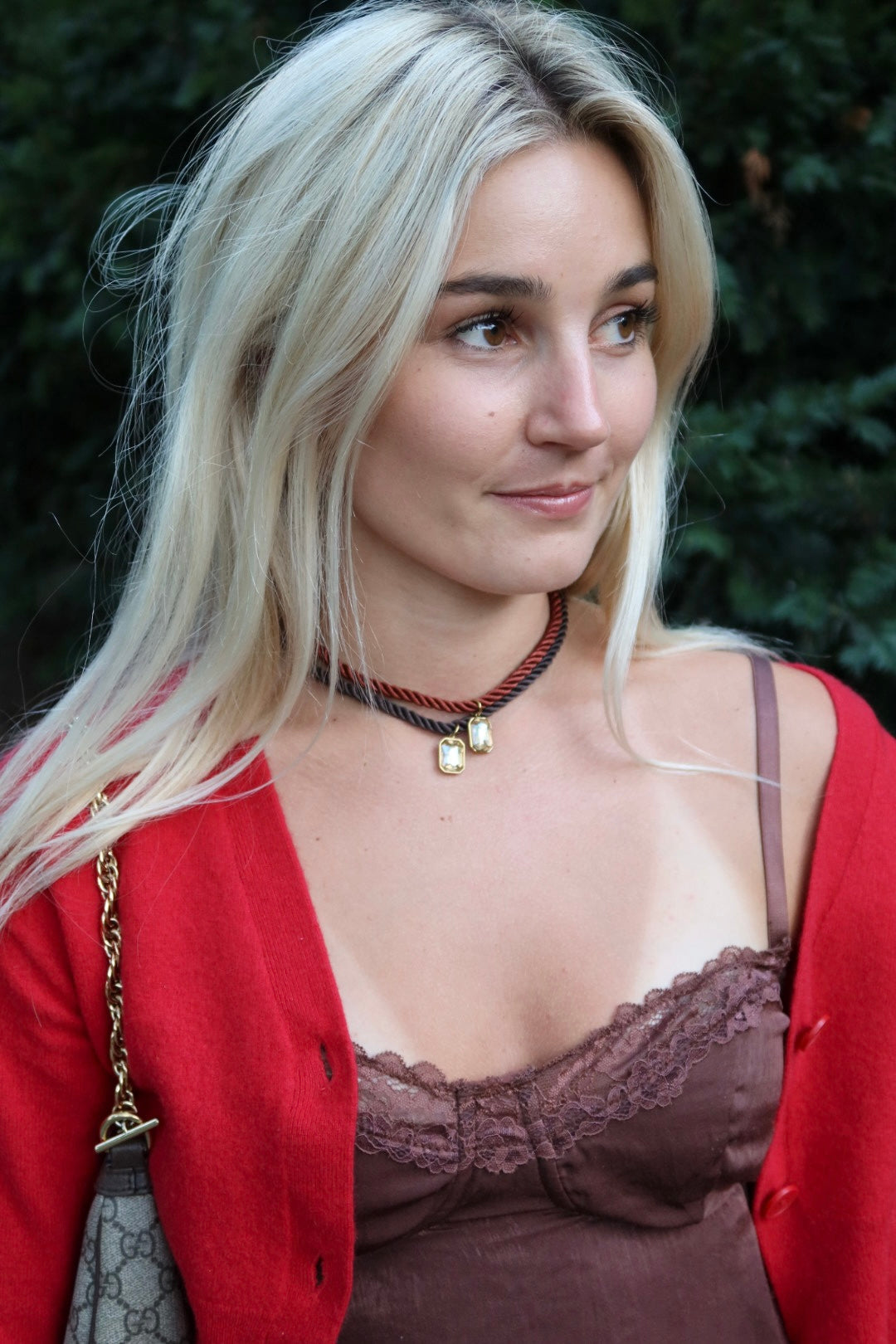 Woman wearing a red cardigan and brown top with a brown rope charm necklace, standing outdoors.