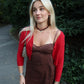 Woman wearing a red cardigan over a brown dress with a necklace, standing outdoors.