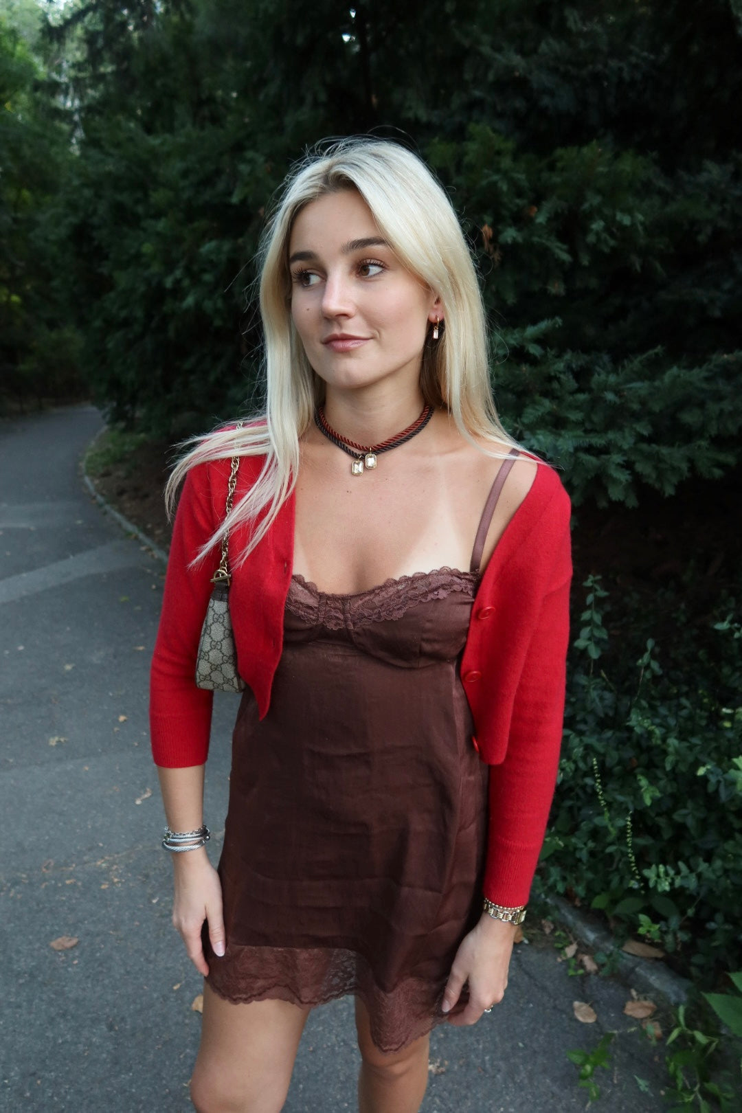 Woman wearing a red cardigan over a brown dress with a necklace, standing outdoors.